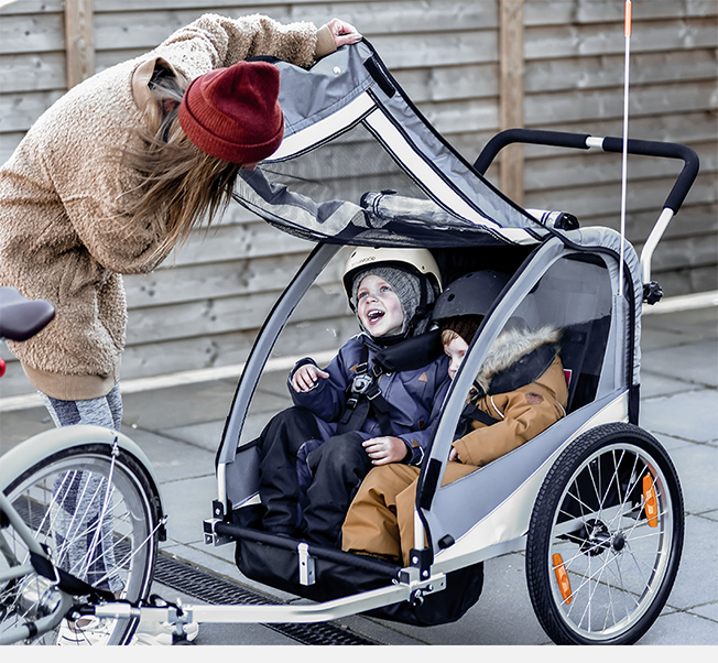 To barn i hjelmer sitter i en sykkelvogn mens en person justerer trekket.