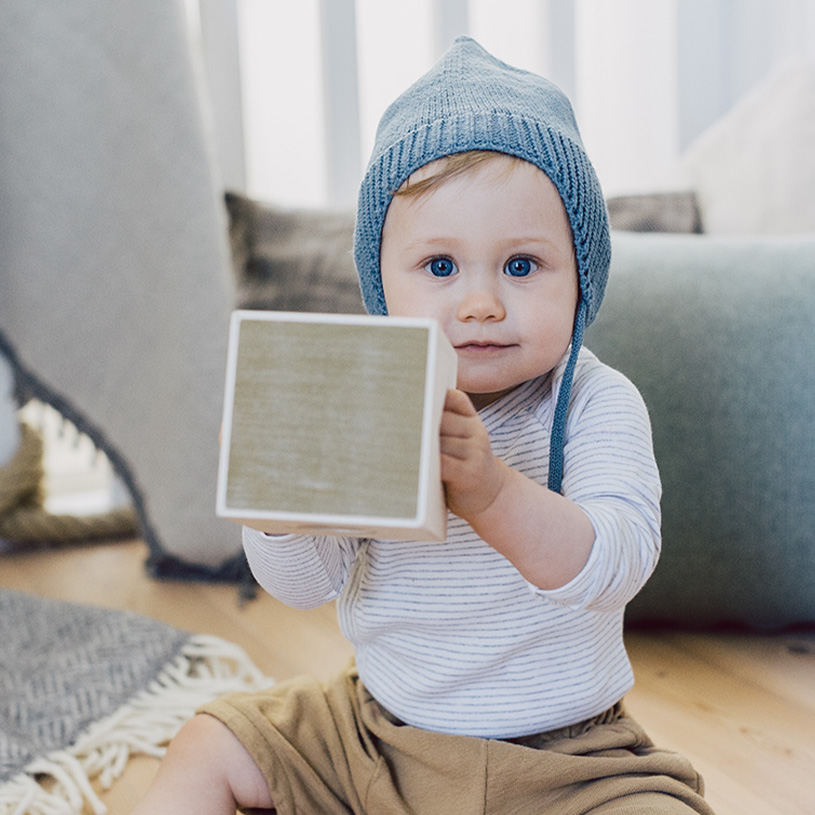 Baby med blå lue holder en trekloss innendørs.