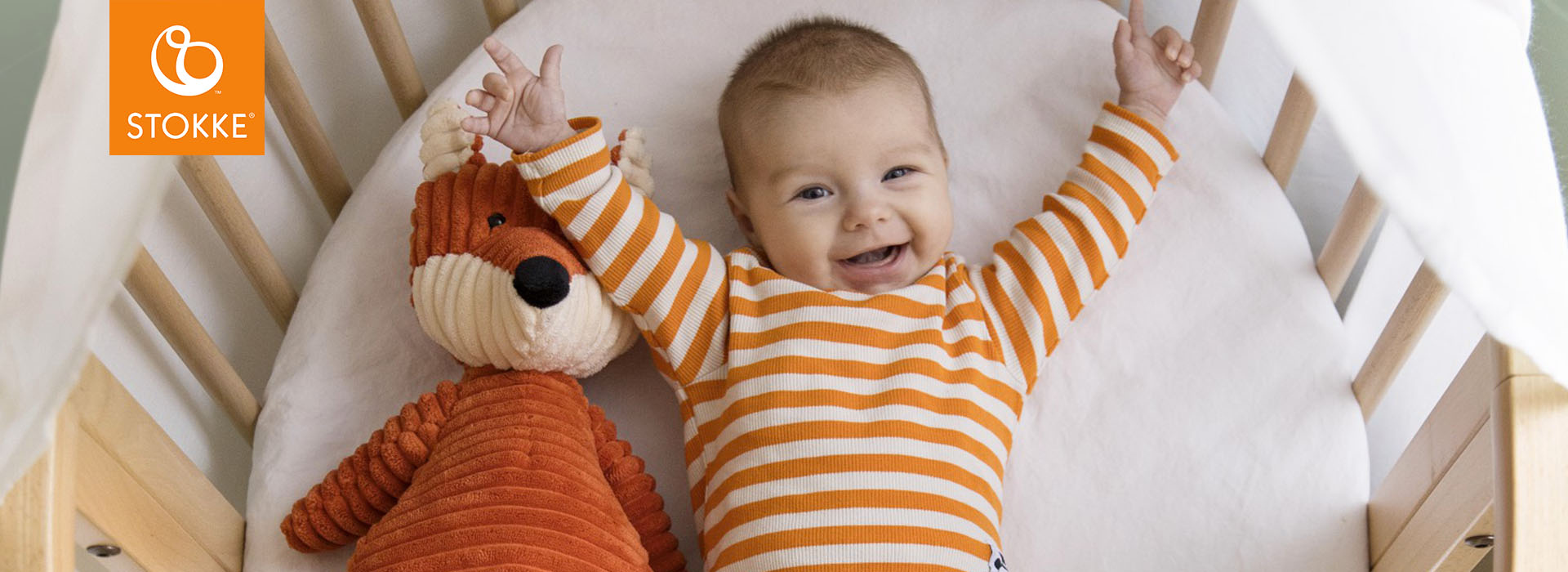 Smilende baby i stripete genser med kosedyr i sengen.