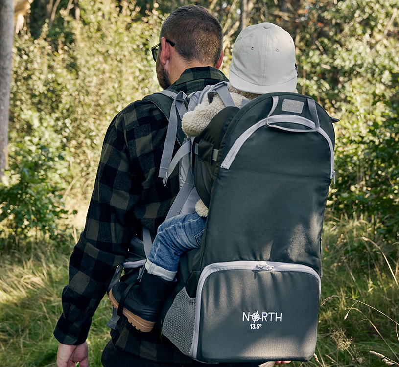 Mann bærer barn i bærestol gjennom skogen.
