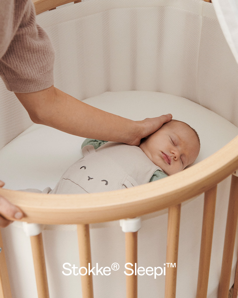 Baby sover i rund sprinkelseng, hånd på pannen.