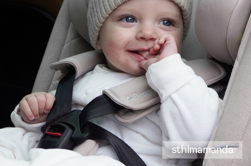 Baby i bilsete med beige lue smiler og suger på fingeren.