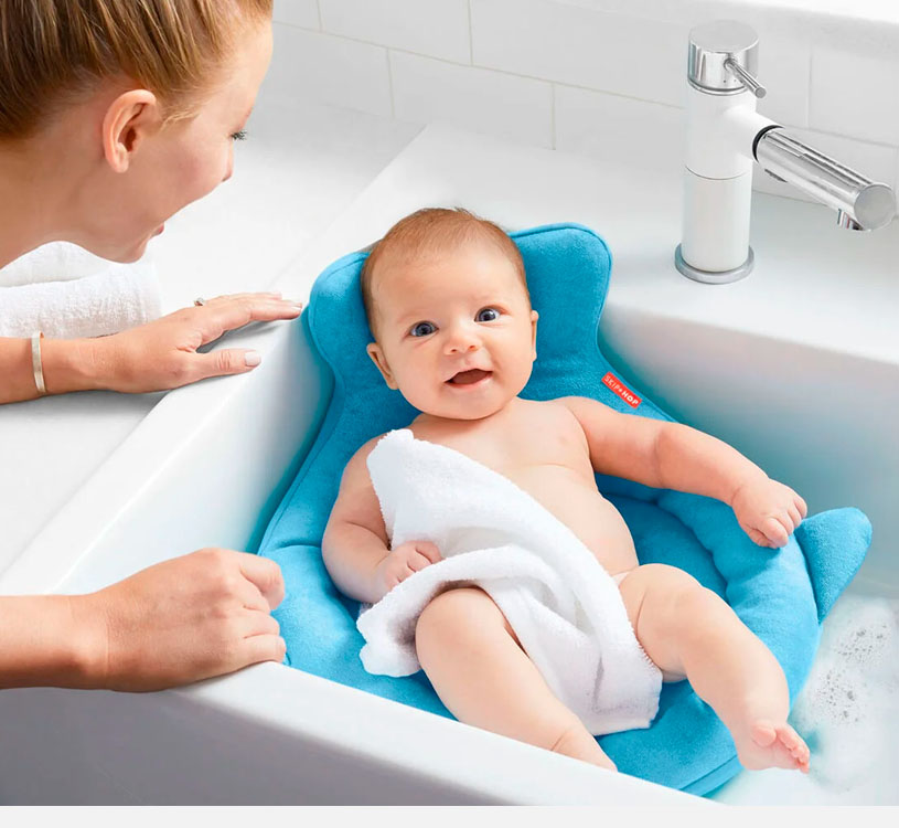 Baby i badekar med blå støttekile, smilende og dekket av håndkle.