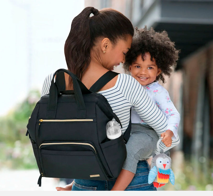 Kvinne med ryggsekk bærer smilende barn med uglebamse.