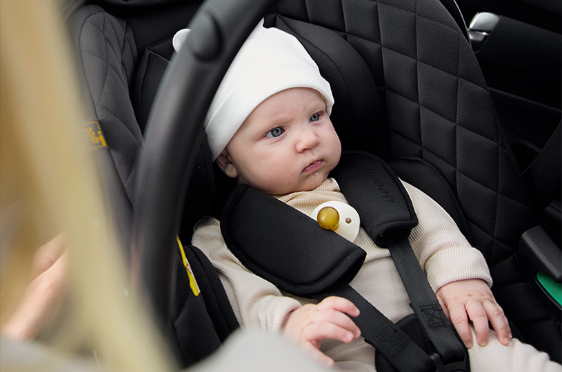 En baby med hvit lue sitter i en bilbarnestol.