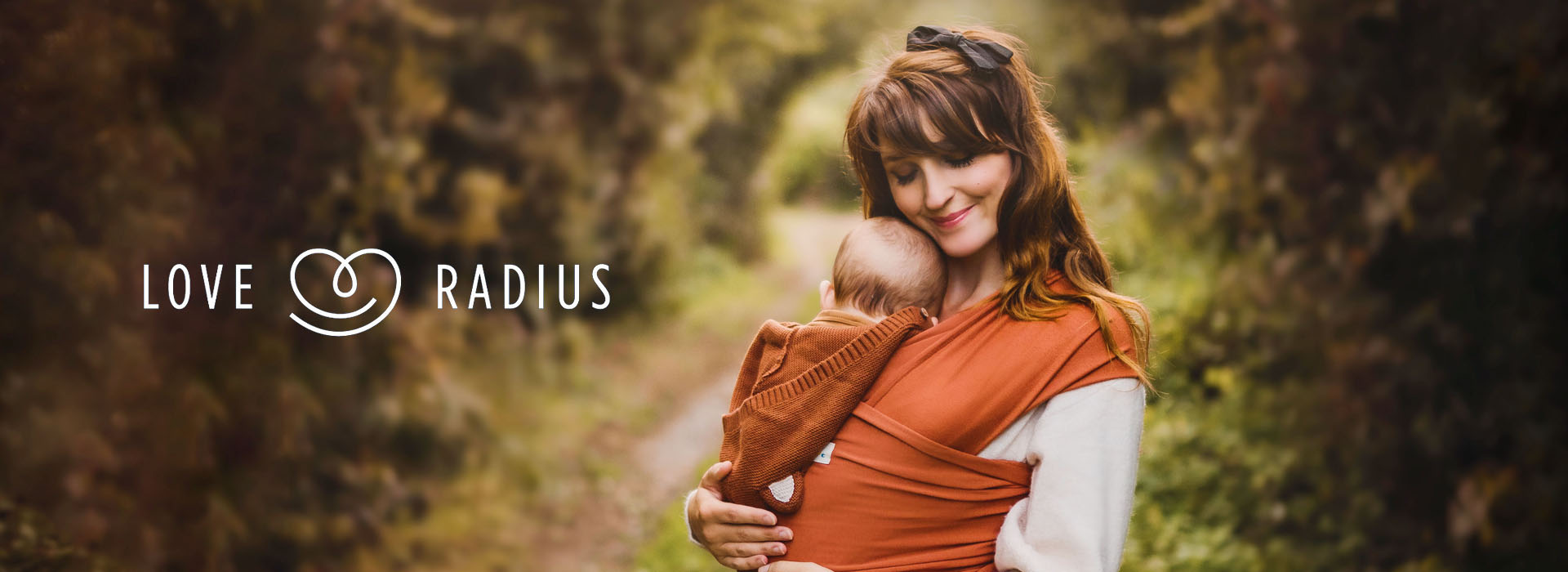 Kvinne holder baby i sjal i skogsområde, logo til venstre.