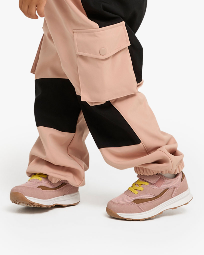 Barneføtter i rosa joggesko og grønne bukser på en gressplen.