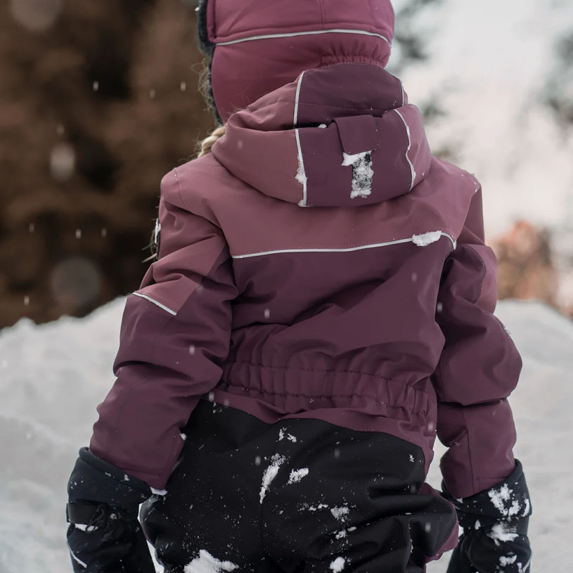 Barn i vinterklær sitter i snøen ved et tregjerde.