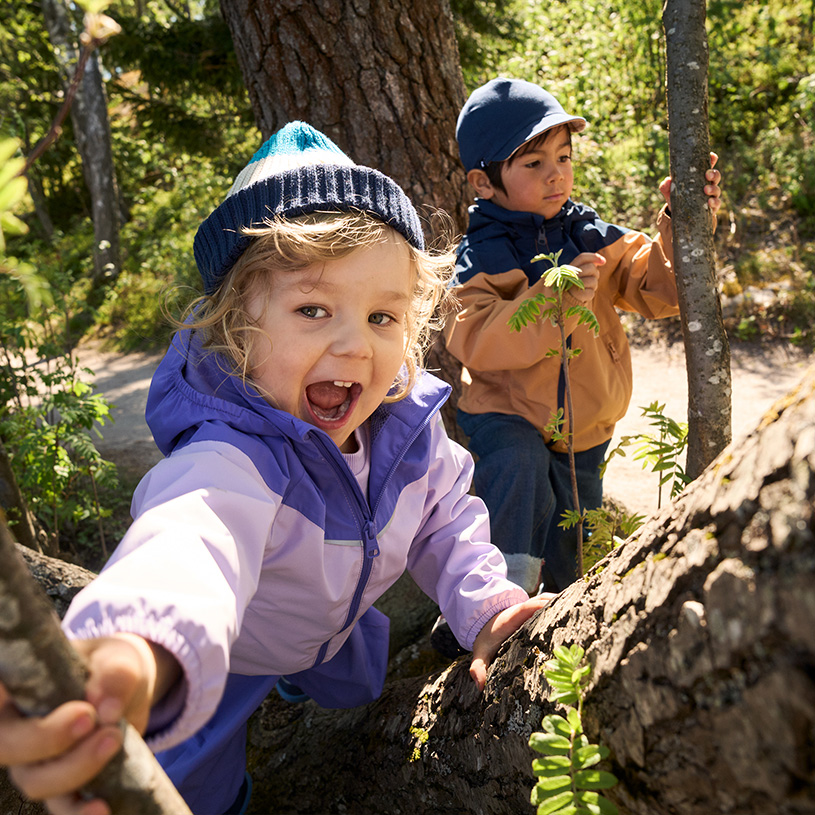To barn i fargerike jakker ser nysgjerrig rundt et tre i skogen.