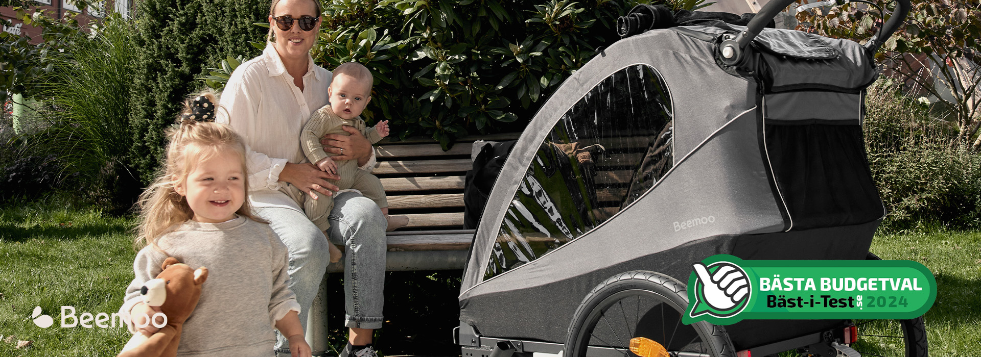 Kvinne og to barn ved benk med sykkelvogn fra Beemoo, smilende i solskinn.