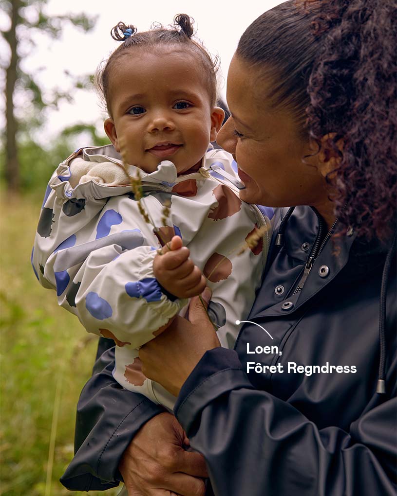 Kvinne holder en smilende baby i en prikkete regnjakke utendørs.