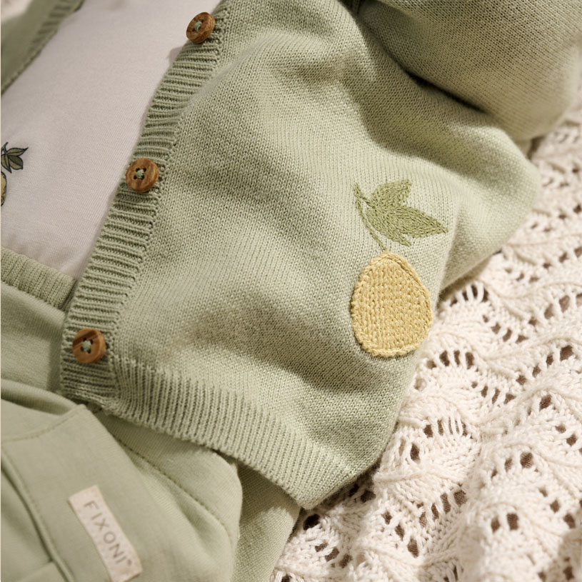 Grønn babyjakke med pærebord og treknapper på hvitt teppe.
