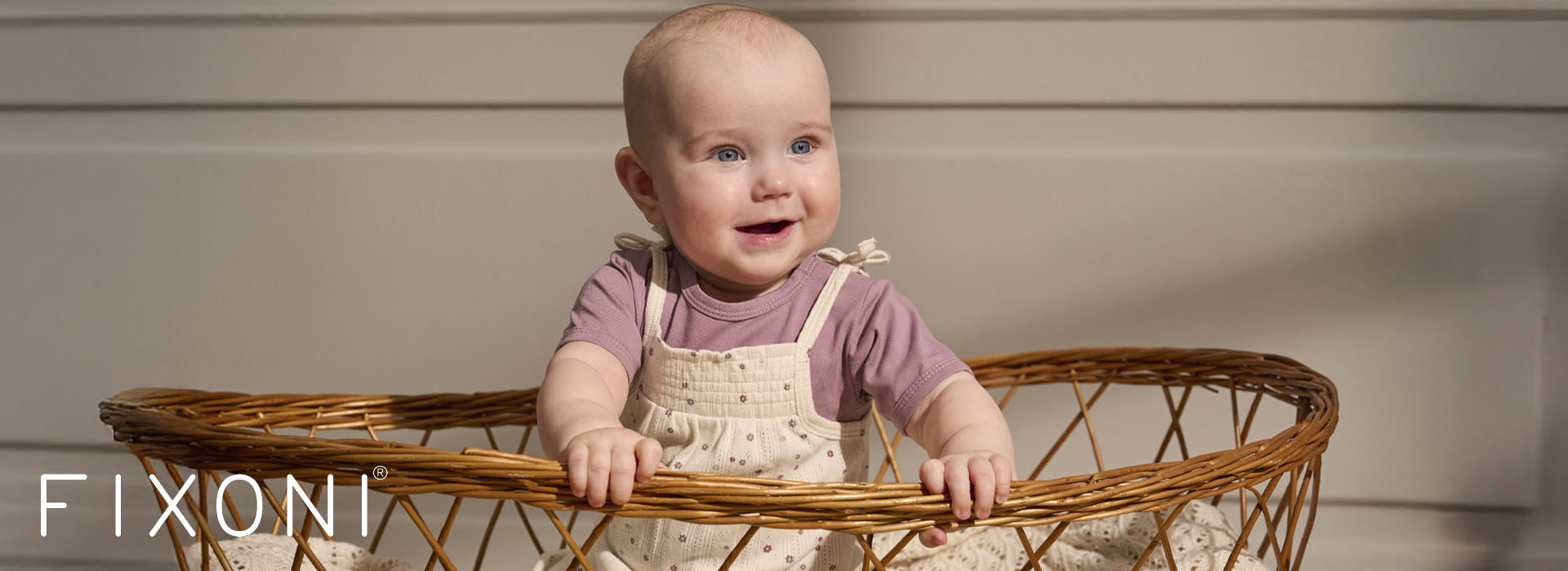 Baby smiler i kurv, kledd i lilla trøye og hvit overall.