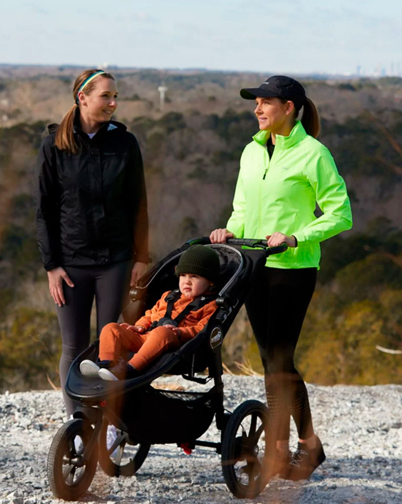 To kvinner med barnevogn på en grusvei med utsikt over en skog.