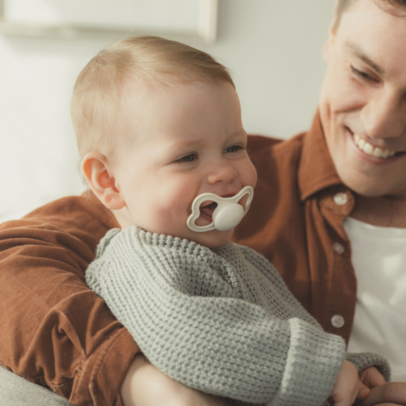 Baby med smokk i armene på en smilende voksen.