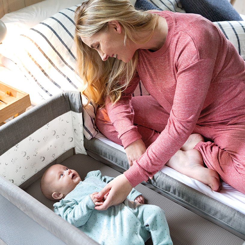 Baby i vippestol med leker, jente sitter ved bord i bakgrunnen.