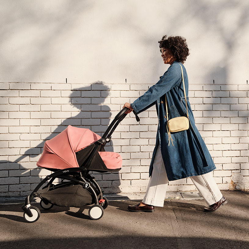 Kvinne i blå frakk går med rosa barnevogn foran hvit murvegg.