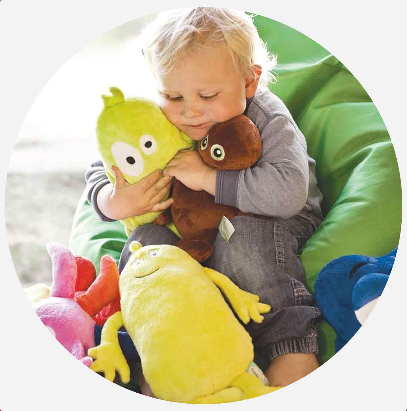 Lite barn klemmer to fargerike kosedyr og sitter i en grønn sakkosekk.