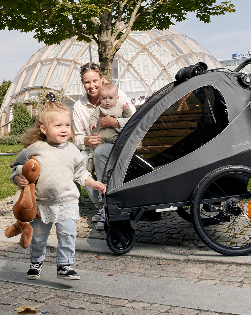 Mor med to barn ved en barnevogn foran et stort glasshus.